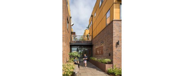 Exterior view of a contemporary multi-storey brick building forming a narrow pedestrian courtyard or entry passage. The passage has paved flooring, raised planted beds, glazed doors, an overhead connecting bridge, and warm timber-toned upper cladding with large windows.
