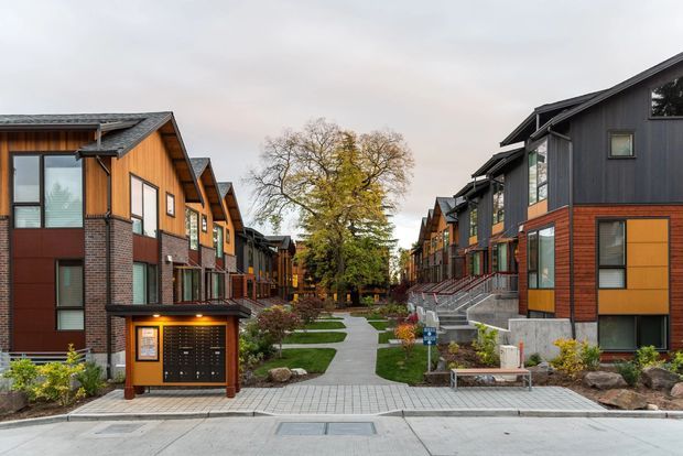 A landscaped pedestrian courtyard running through a contemporary residential townhouse development. Repeated multi-storey homes line both sides, combining dark vertical cladding, warm timber-toned panels, red-brown brick, and large windows beneath steeply pitched gable roofs. The central walkway includes paving, lawns, planting, benches, lighting, and steps, creating a cohesive shared outdoor space.