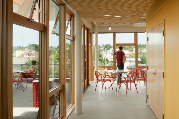 A bright, contemporary communal dining or breakout area with extensive timber-framed glazing along one side and at the far end, opening onto a balcony or terrace. The space features red wire-frame café chairs, small round tables, a smooth pale concrete floor, a warm timber slatted ceiling, white cylindrical columns, and light wood wall cabinetry or doors. A person stands near the far windows, with an urban treetop outlook beyond.