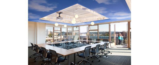 A bright contemporary commercial meeting and conference room with a large U-shaped arrangement of tables and black ergonomic office chairs. The room has extensive floor-to-ceiling glazing, timber-framed glass doors and windows, a white coffered ceiling with suspended globe lights and a ceiling-mounted projector. An outdoor terrace and urban surroundings are visible beyond the glazing.