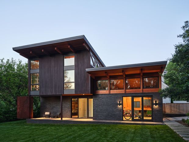 Exterior view of a contemporary two-level residence with dark vertical timber cladding, charcoal brickwork, extensive glazing, and broad low-pitched roof planes. Warm interior lighting is visible through the windows and glazed doors, with a lawn, paved walkway, and mature landscaping around the house.