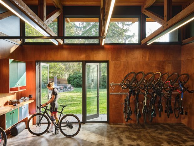A warm, timber-lined bicycle storage and mudroom space with a polished concrete floor, extensive exposed timber framing, high-level glazing, and large opening doors to a landscaped garden. Multiple bicycles are mounted on the wall, while colourful built-in cabinetry and a long linear LED light add functional contemporary detailing.