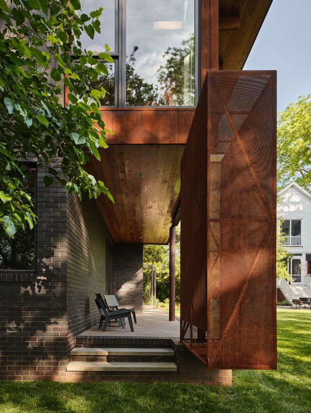 Exterior view of a contemporary residential entry and covered porch. The design features dark brick walls, a warm timber soffit, large glazed openings, and a tall rusted steel privacy or screen door. Brick steps lead from the lawn to the sheltered entry, with mature planting and a neighbouring traditional house visible in the background.