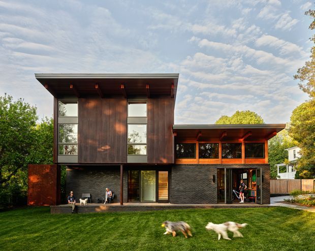A contemporary two-storey residential house set within a leafy suburban landscape. The home features a strongly articulated flat roof with deep overhangs, dark vertical timber cladding on the upper level, dark brick masonry at ground level, extensive glazing, and warm timber-framed outdoor living areas. A broad lawn occupies the foreground, with mature trees and neighbouring homes visible around the site.