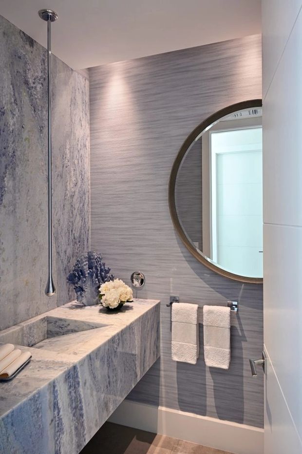 A refined contemporary bathroom with a sculptural freestanding-style stone basin and integrated ledge, vertically veined grey stone wall cladding, and a horizontally textured grey feature wall. A large round dark-framed mirror, wall-mounted chrome tapware, twin folded towels, recessed lighting, and warm stone-look floor tiles complete the compact space.