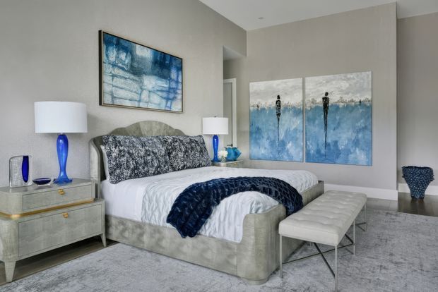 A refined bedroom with a large upholstered bed, white bedding, and a deep blue textured throw. The room features coordinated blue-and-white abstract artwork, blue glass lamps and accessories, a mirrored bedside cabinet, a padded bench at the foot of the bed, pale carpet, and neutral textured walls. The overall look is polished, calm, and contemporary with a strong blue accent palette.