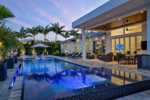 Twilight view of a luxurious modern resort-style residence with a large illuminated swimming pool and raised spa in the foreground. The open-sided covered outdoor living area includes lounge seating, a dining setting, ceiling fans, recessed lighting, and a warm timber-lined ceiling. Floor-to-ceiling glazing opens toward the pool, with tropical palms and landscaped planting surrounding the property.