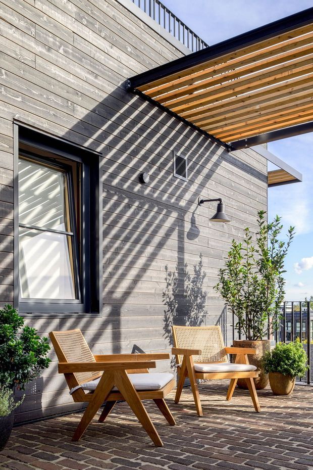 A bright rooftop or elevated outdoor terrace beside a contemporary building, featuring grey horizontal weatherboard cladding, black-framed glazing, a timber pergola with open slatted roof, brick-paved flooring, black metal balcony railing, outdoor wall lighting, and two timber lounge chairs with potted greenery. Strong sunlight creates shadows from the pergola across the facade and terrace.