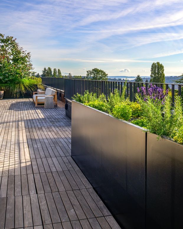 A spacious rooftop or elevated outdoor terrace with dark timber decking, black metal privacy balustrades, and large integrated planter boxes filled with ornamental grasses and flowering plants. Minimal outdoor lounge chairs sit at the far end, with an expansive blue-water and tree-lined view beyond. The scene is bright, natural, and contemporary, with strong linear decking and planter geometry.
