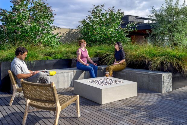 A landscaped outdoor terrace with timber decking, built-in concrete bench seating, and a square concrete fire pit table. Three people sit around the seating area, which is integrated into raised garden beds with ornamental grasses and mature planting. A partially visible dark-clad building forms the background.