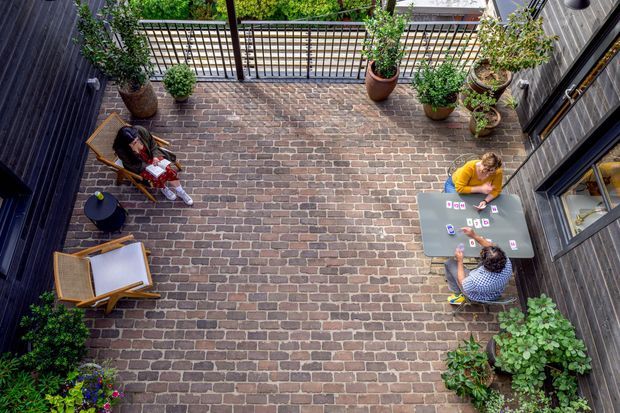Top-down view of a compact enclosed courtyard between dark-clad building walls. The space has a warm brick-paved surface, black metal railing and trellis, abundant potted greenery, timber lounge chairs, a small side table, and a table where two people are playing cards. Large windows and doors open onto the courtyard from the surrounding building.