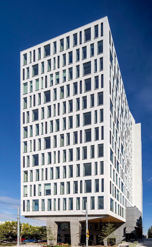 Exterior photograph of a tall contemporary multi-storey commercial or office building on an urban corner site. The building has a crisp white rectilinear facade with a dense, varied grid of narrow and broad recessed windows, a projecting upper volume, and a darker grey stone or concrete base with a covered ground-level entry. Clear blue sky and surrounding streetscape reinforce the urban setting.