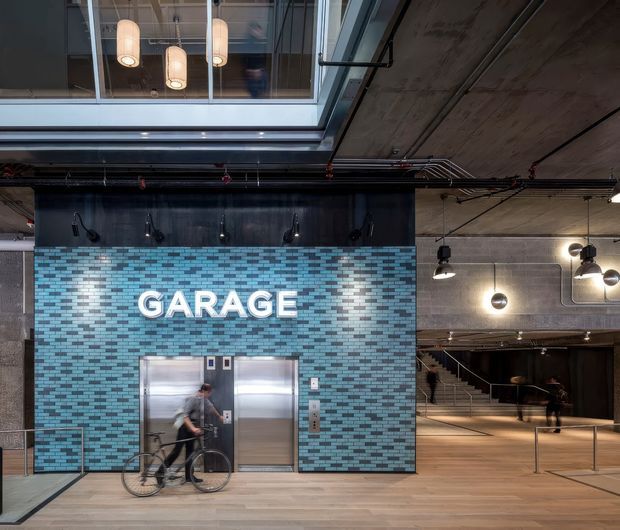 Interior view of a large contemporary commercial space with an industrial aesthetic. A blue and grey mosaic-tile wall marked “GARAGE” contains stainless-steel elevator doors, while exposed concrete ceilings, visible services, black metal detailing, pendant lights, and stairs reinforce the raw warehouse-like character. A mezzanine or glazed upper level is visible above.