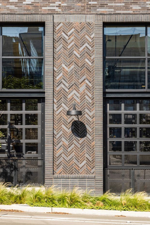Close-up exterior view of a contemporary multi-storey brick-clad building facade. The elevation features dark-framed glazed windows and doors, a prominent central vertical panel of patterned herringbone brickwork, an exterior wall light, metal mesh balcony or guardrail panels, and landscaped grasses along the street edge.