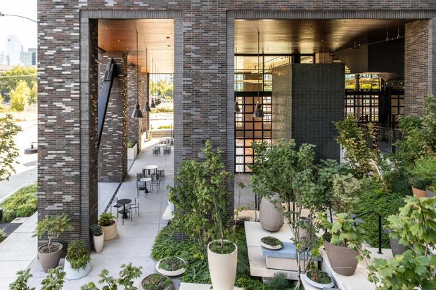 Exterior architectural view of a contemporary multi-storey building with a dark variegated brick facade, large open glazed bays, and warm timber soffits. A landscaped terrace with abundant potted greenery, outdoor café-style tables and chairs, and a raised seating area occupies the foreground. Interior spaces are visible through the glazing, including a warm-toned lobby or dining area.