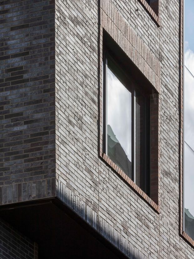 Close-up exterior architectural view of a contemporary brick facade, showing textured dark and pale brickwork, deep-set tall windows with dark frames, and strong geometric projecting and recessed wall forms. The image emphasizes material texture, shadow, and facade detailing rather than the overall building.