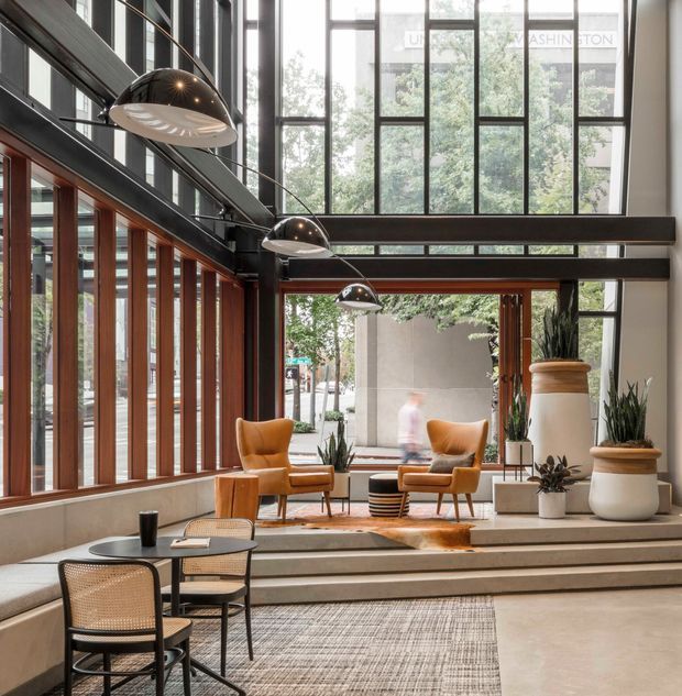 A sophisticated double-height commercial lobby or lounge with extensive floor-to-ceiling glazing, dark exposed steel beams, warm timber-framed windows, and stepped concrete or stone platforms. The space includes caramel leather lounge chairs, small round tables, woven cane chairs, large planters, pendant lighting, and layered rugs, creating a refined, welcoming atmosphere with strong indoor-outdoor visual connection.