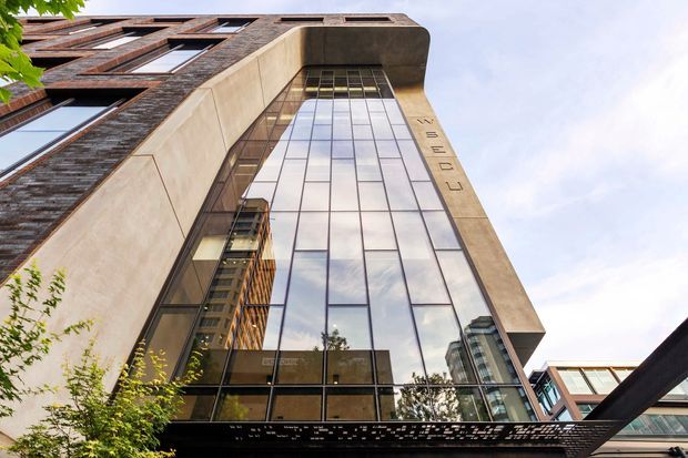Upward-looking exterior view of a contemporary multi-storey commercial building. The facade combines extensive floor-to-ceiling curtain-wall glazing, pale concrete or stone cladding, and dark textured masonry panels. A tall recessed glazed entrance or atrium is framed by a deep projecting canopy, with neighboring urban buildings reflected in the glass.
