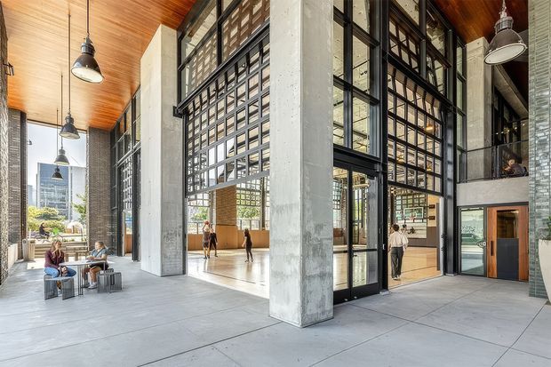A spacious contemporary commercial interior with a high timber-lined ceiling, exposed concrete columns, polished concrete flooring, and extensive black metal-framed glazing. Large industrial-style grid windows and glazed doors create an airy atrium or lobby, with pendant lights, a small seating area, and people moving through the space. Urban buildings are visible beyond the glazing.