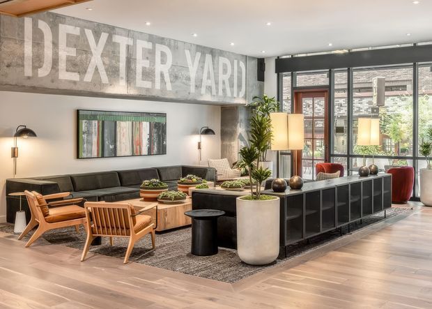 A spacious, stylish living room with an industrial-inspired interior. The room features a large black modular sofa, mid-century-style timber lounge chairs, a low timber coffee table, a long black built-in console or room divider, pendant and floor lamps, indoor plants, and a large distressed wall sign reading “DEXTER YARD.” Floor-to-ceiling black-framed glazing opens visually toward an outdoor courtyard or terrace. Warm timber flooring, concrete-look wall finishes, and layered lighting create a refined urban warehouse atmosphere.