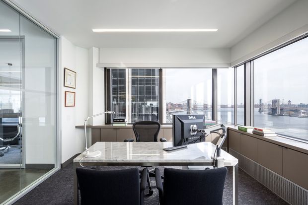 Bright contemporary office overlooking a waterfront cityscape, with a marble-topped meeting desk, two dark upholstered visitor chairs, an ergonomic desk chair, dual computer monitors, task lighting, and extensive floor-to-ceiling glazing. A glass-partitioned room is visible to the left, while built-in dark cabinetry runs beneath the windows.