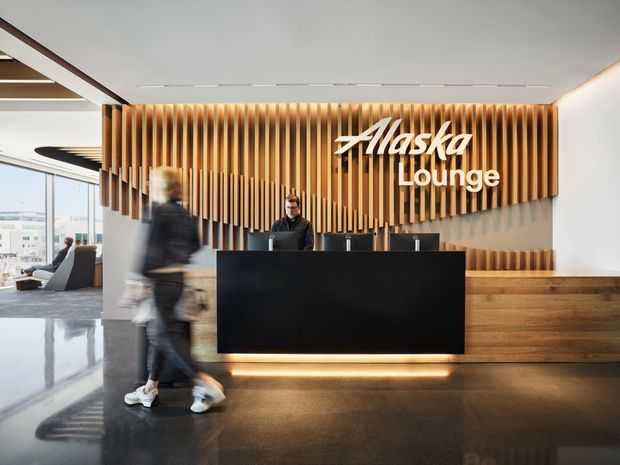 A modern airport lounge reception area with a large black reception desk, warm timber-clad feature wall with vertical slats, illuminated signage reading “Alaska Lounge,” polished dark flooring, and expansive glazing on the left. A staff member stands behind the desk while a blurred visitor walks through the foreground.