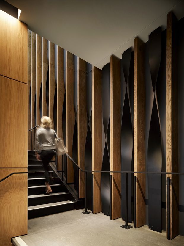 A dramatic, contemporary stairway with black steps and slim black metal balustrades, set beside a sculptural wall of alternating vertical timber fins and dark panels. Warm concealed lighting highlights the timber grain, while a pale concrete-like floor and ceiling provide contrast. A person is ascending the stairs.