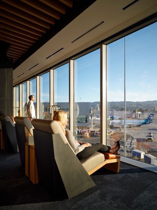 A refined airport lounge or terminal waiting area with floor-to-ceiling glazing overlooking an active airport apron and aircraft. The interior features sculptural upholstered lounge chairs with timber frames, dark carpet, warm timber ceiling beams, integrated linear lighting, and a calm, high-end hospitality atmosphere.