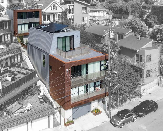 Aerial view of a compact contemporary multi-storey urban residence with a sharply folded metal-clad upper volume, warm timber-toned facade panels, extensive glazing, roof terrace, rooftop solar panels, and balconies. The home sits within a dense leafy neighbourhood of mixed residential buildings, with overhead utility wires and street parking visible.