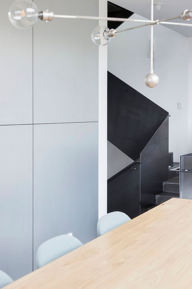 A bright, minimalist dining area with a large pale timber dining table and pale upholstered chairs in the foreground. A sculptural contemporary pendant with spherical bulbs hangs overhead. Behind the table is a sharply geometric black metal staircase with grey metal treads, set against white walls and large pale built-in wall panels.