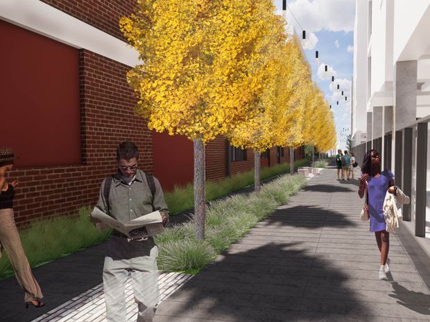 A rendered urban pedestrian streetscape beside a contemporary multi-storey building. The walkway is lined with yellow-leaved trees, ornamental grasses, and hanging lights, with a dark red brick facade and large red wall panels on the left and a white glazed building frontage on the right. Several pedestrians occupy the pathway.