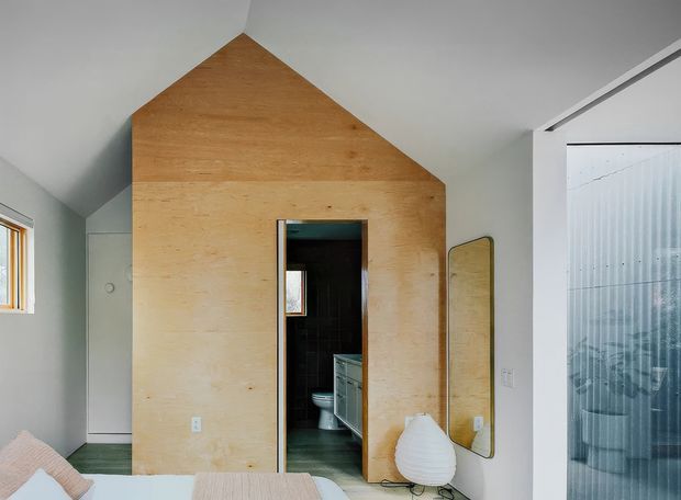 A compact contemporary bedroom with a distinctive plywood-clad, house-shaped partition enclosing a dark tiled bathroom. The room has a sloping ceiling, pale walls, a light timber or plywood floor, a small window, a full-height rounded mirror, and a translucent ribbed-glass sliding door or screen. A bed with pale bedding and a sculptural white chair are partially visible in the foreground.