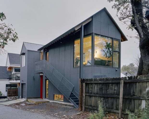 Exterior view of a compact two-storey dwelling with a steep gabled roof and dark corrugated metal cladding. The building features prominent floor-to-ceiling corner glazing, an external steel staircase and balcony, and a bright red entry door. A weathered timber boundary fence and mature tree frame the foreground, with neighbouring residential buildings visible behind.