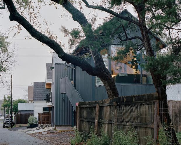 Street-level exterior view of a contemporary multi-storey residential building, partly concealed by mature trees and a timber fence. The building features dark grey corrugated metal cladding, angular roof forms, projecting upper-level glazing, and an external stair enclosure. A narrow urban residential lane and neighbouring homes are visible.