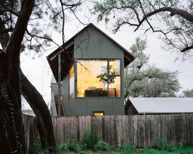 Exterior view of a compact, narrow-gabled dwelling set behind a weathered timber fence and mature trees. The building has dark grey-green vertical corrugated cladding, a steep pitched gable roof, large framed windows glowing warmly from the interior, and a small lower-level window. A secondary low corrugated-roof structure is visible behind the fence.