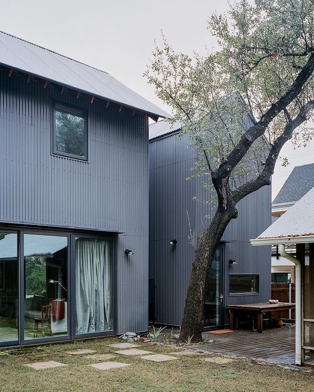 Exterior view of a contemporary multi-volume residential building arranged around a landscaped courtyard. The dark grey corrugated metal-clad walls and pitched metal roofs frame large glazed doors, a timber deck, and an established tree. A covered outdoor dining or work area is visible on the right, with stepping stones leading through the lawn.