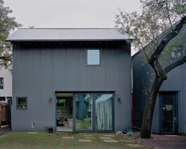 Exterior view of a compact contemporary two-storey residential building clad in dark grey vertically ribbed metal, with a light grey corrugated metal roof, large ground-floor glazed doors, and a smaller upper window. A mature tree, stepping-stone path, and timber deck sit in the foreground, while an adjoining similarly clad volume is visible to the right.