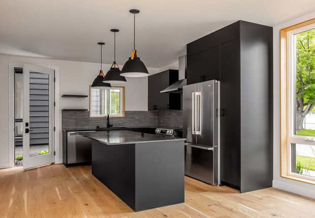 A contemporary black kitchen with a central island, matte dark cabinetry, stainless-steel refrigerator and appliances, grey horizontal-tile backsplash, timber flooring, and multiple black pendant lights. Large windows and a glazed door bring in natural light and views of greenery.