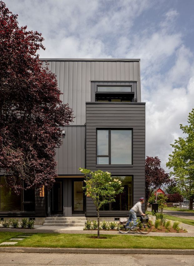 Street-facing contemporary multi-storey townhouse with a compact, vertically composed facade. The building combines dark horizontal cladding on the projecting lower volume with light grey corrugated metal cladding above, large rectangular windows, a recessed entry, and a small front garden. A cyclist passes in the foreground, with neighbouring homes and mature trees visible along the suburban street.