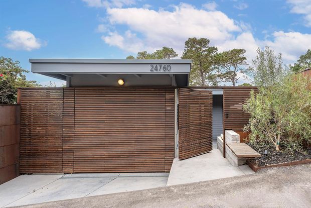 Street-level view of a compact contemporary residential entrance and garage frontage. The facade is screened with horizontal dark timber battens, including a broad garage door and a pedestrian gate, beneath a shallow projecting flat roof. A concrete driveway and pathway lead to the entry, with built-in concrete seating and sparse landscaping at the side. The address number is mounted on the roof fascia.