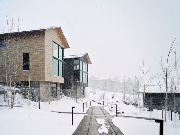 Snow-covered modern building complex viewed from a paved pedestrian path. The buildings feature angular pitched roof forms, timber-clad upper volumes, dark lower sections, exposed concrete bases, and large floor-to-ceiling glazing. Leafless trees, snow, and minimalist black path railings create a stark alpine or cold-climate setting.