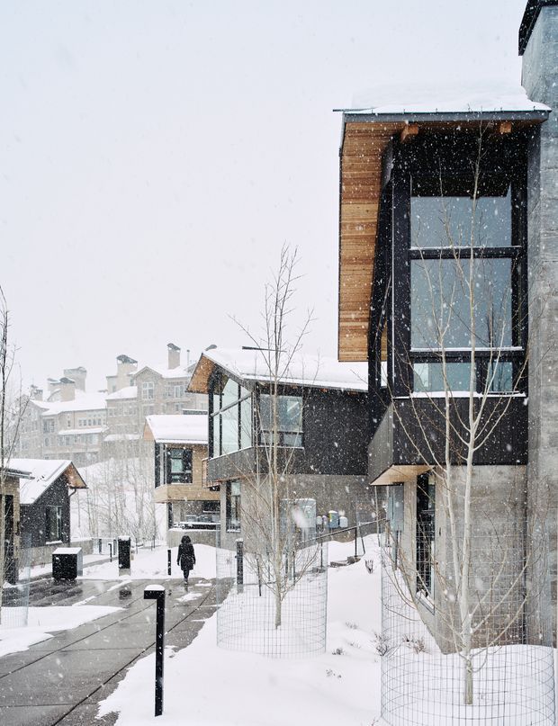 Snow-covered contemporary residential development viewed from a pedestrian walkway. The foreground building combines dark cladding, warm horizontal timber cladding, large black-framed glazing, and exposed grey masonry or concrete. Similar multi-storey buildings and steep snowy roofs recede into the background, with young leafless trees and bollard lighting along the path.