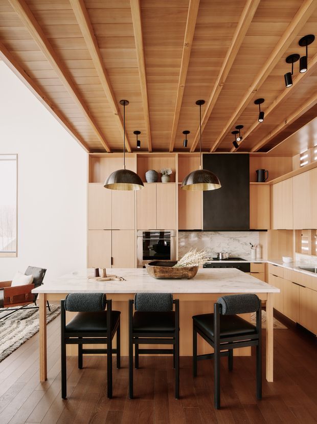 Warm, refined open-plan kitchen and dining area with extensive light timber cabinetry and a dramatic exposed timber-lined ceiling with beams. A large pale stone island doubles as a dining table with three black upholstered chairs. The kitchen includes integrated appliances, a dark feature splashback or wall panel, white veined stone benchtops, and contemporary black pendant and track lighting. A living area is partially visible at left.