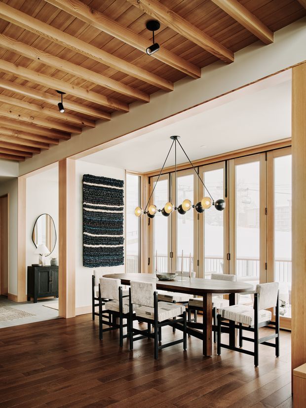 A warm, contemporary dining area with a long dark timber dining table surrounded by upholstered dining chairs. The room features a dramatic exposed timber beam ceiling with timber planks, dark timber flooring, pale walls, large timber-framed glazed folding or sliding doors, and a sculptural linear pendant light with globe bulbs. An adjacent living or entry area is visible to the left, with a console, mirror, and textured wall hanging.