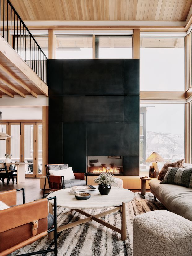 A sophisticated double-height living room with a dramatic black fireplace wall, extensive warm timber ceiling and structural framing, and large glazed openings. A mezzanine with black metal balustrading overlooks the space. The room is furnished with a large sofa, leather lounge chairs, a round coffee table, layered rugs, and a visible dining area beyond. A snowy mountainous landscape is visible through the windows.