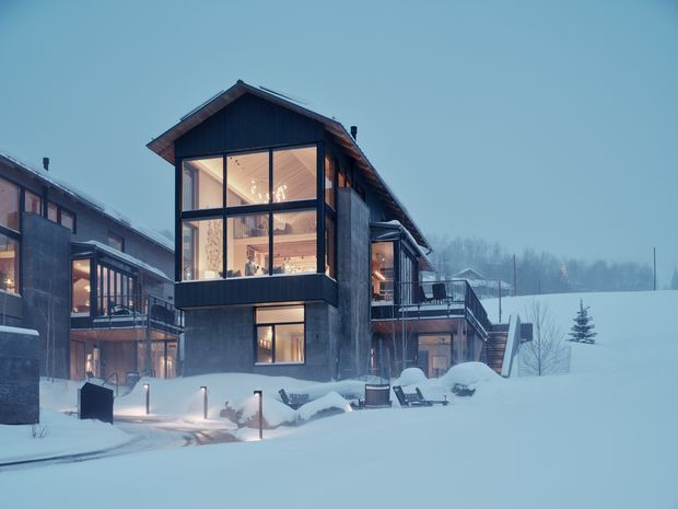 Exterior view of a contemporary multi-level mountain dwelling in a snowy landscape. The building has dark timber or stained cladding, extensive floor-to-ceiling glazing, projecting balconies, and a steep gabled roof with visible eaves. Warm interior lighting contrasts with the cool blue snow and overcast surroundings. Adjacent connected or neighbouring building volumes are visible on the left, with a snow-covered outdoor area and pathway in the foreground.