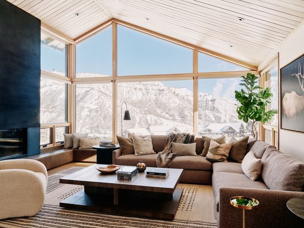 A warm, contemporary living room with a large L-shaped sectional sofa, layered neutral textiles, a low dark wood coffee table, and a sculptural lounge chair. Floor-to-ceiling timber-framed glazing provides expansive views of a snow-covered mountain landscape. The room has a sloped timber-lined ceiling, a dark fireplace wall, recessed downlights, framed artwork, and indoor greenery.