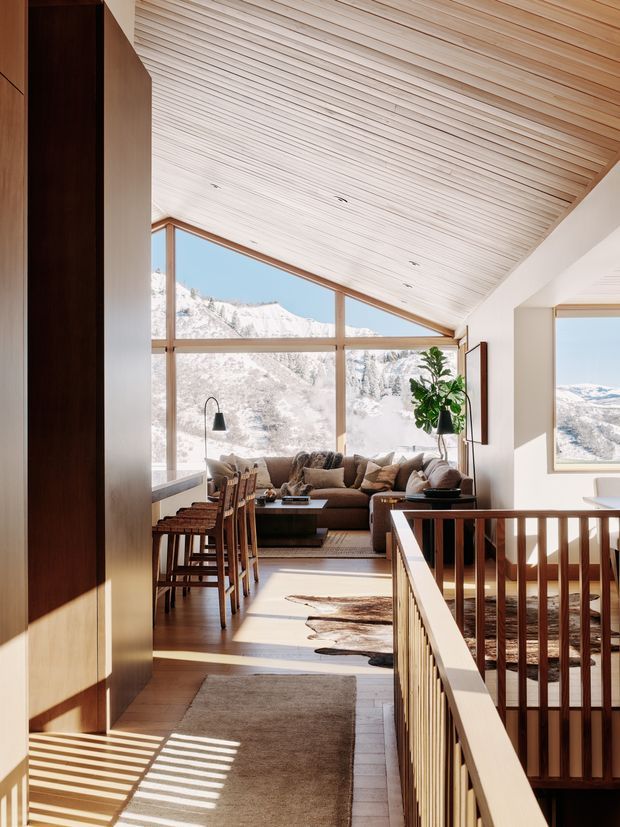 Bright alpine living area viewed from an upper-level landing, with expansive floor-to-ceiling glazing framing snow-covered mountains. The room features a pitched, wood-slat ceiling, warm timber flooring and joinery, a brown sectional sofa, dining counter with timber stools, indoor plant, and timber stair or balustrade in the foreground. Strong natural light creates striped shadows across the floor.
