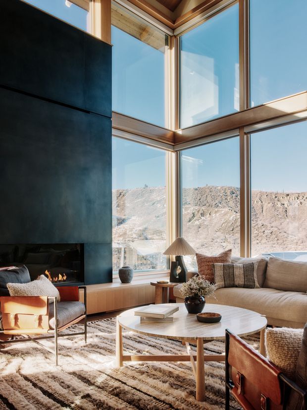 A refined living room with dramatic floor-to-ceiling corner glazing overlooking a rugged mountainous landscape. The room features a tall dark stone or metal fireplace surround with a linear fire, warm timber window framing and ceiling detailing, a large neutral sectional sofa, leather lounge chairs, a round light stone coffee table, and a textured patterned rug. The space is bright, warm, and strongly connected to the landscape.