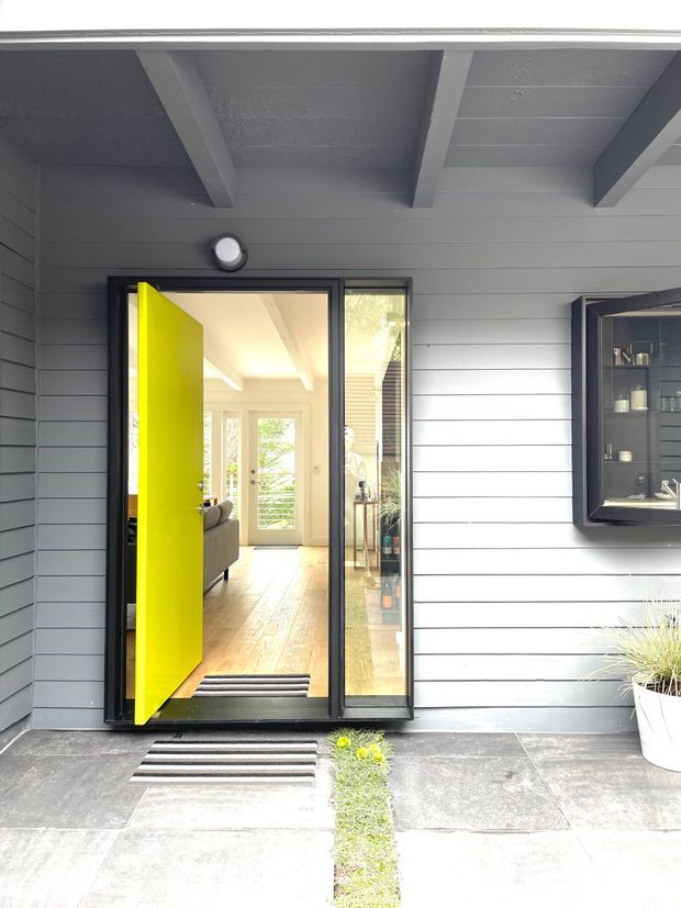 A bright contemporary home entry viewed from the exterior. The facade is clad in horizontal grey weatherboards beneath a covered porch with exposed painted beams. A vivid lime-yellow pivot or hinged front door opens into a light-filled hallway with timber flooring and glazing beyond. Large grey paving slabs, striped doormats, a narrow planted joint, and a wall-mounted dark display cabinet complete the entry.
